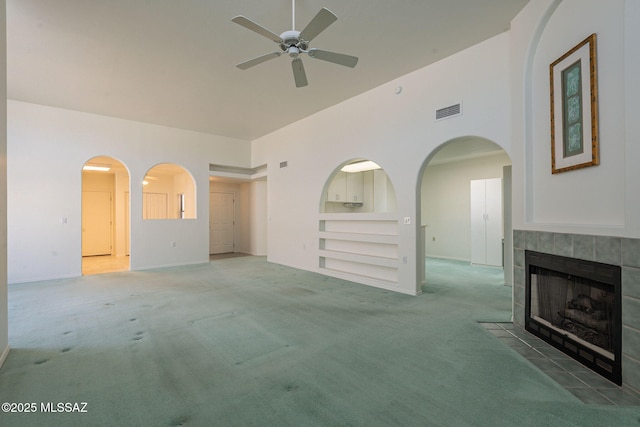 unfurnished living room with built in features, ceiling fan, a towering ceiling, carpet flooring, and a tiled fireplace