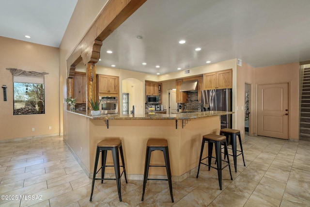 kitchen with stone counters, backsplash, stainless steel appliances, a kitchen bar, and wall chimney exhaust hood