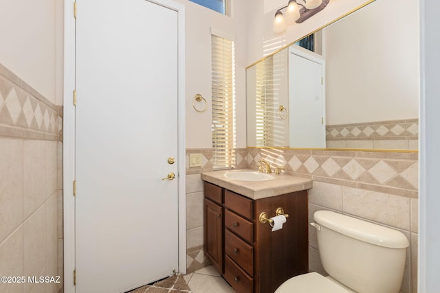 half bathroom featuring tile patterned floors, toilet, tile walls, wainscoting, and vanity