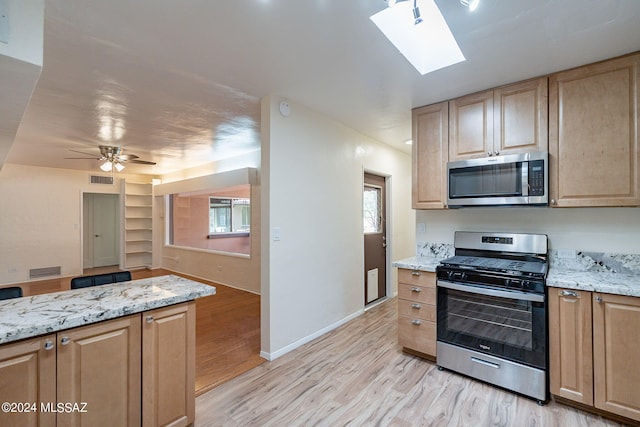 kitchen featuring ceiling fan, a skylight, stainless steel appliances, light stone countertops, and light wood-type flooring