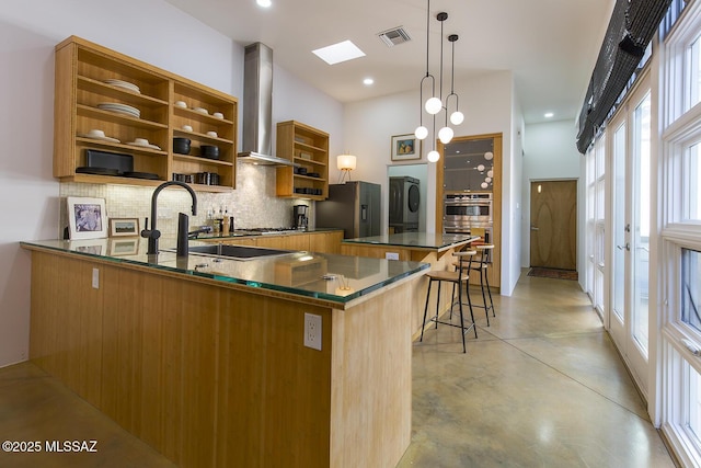 kitchen featuring pendant lighting, wall chimney range hood, backsplash, stainless steel appliances, and an island with sink