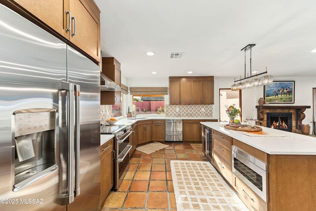 kitchen featuring wall chimney exhaust hood, sink, built in appliances, hanging light fixtures, and decorative backsplash