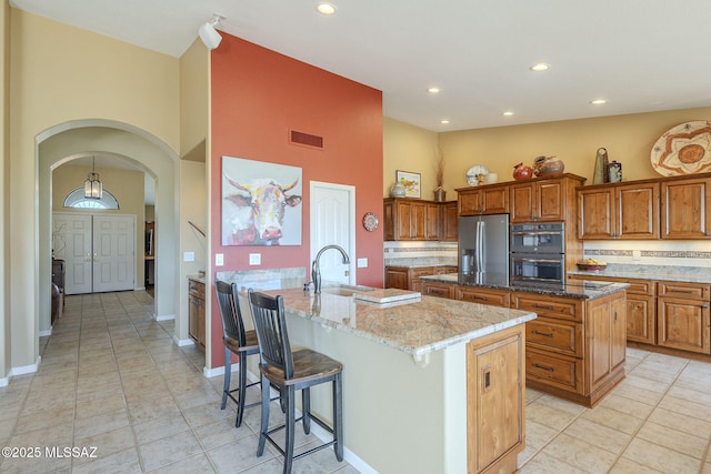 kitchen with tasteful backsplash, brown cabinetry, a sink, an island with sink, and stainless steel fridge