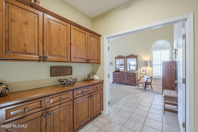 kitchen featuring baseboards, brown cabinetry, light tile patterned flooring, and light colored carpet