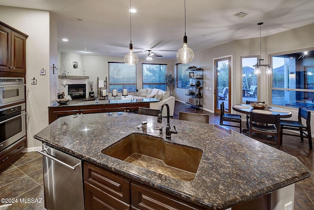 kitchen featuring appliances with stainless steel finishes, an island with sink, sink, dark stone counters, and hanging light fixtures