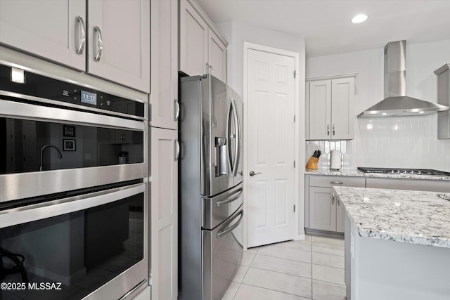 kitchen with gray cabinetry, decorative backsplash, light tile patterned floors, stainless steel appliances, and wall chimney range hood