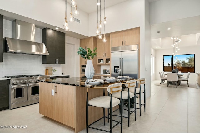 kitchen featuring dark stone counters, hanging light fixtures, high end appliances, a center island, and wall chimney range hood