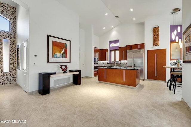 kitchen featuring dark countertops, visible vents, an island with sink, high vaulted ceiling, and built in refrigerator