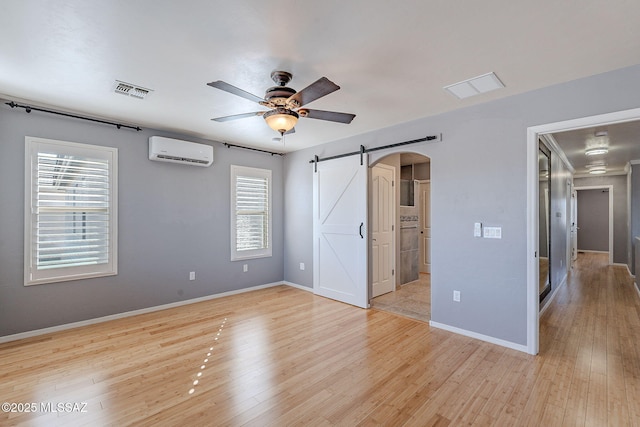 unfurnished bedroom with ceiling fan, a barn door, a wall mounted AC, and light wood-type flooring