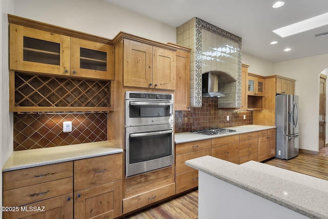 kitchen with tasteful backsplash, light stone counters, appliances with stainless steel finishes, light hardwood / wood-style floors, and wall chimney range hood