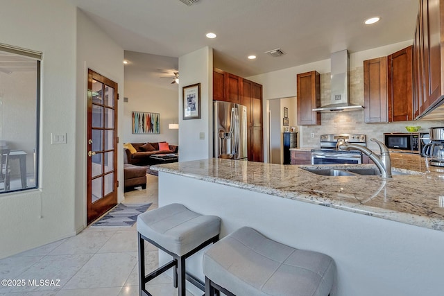 kitchen featuring wall chimney range hood, kitchen peninsula, stainless steel appliances, light stone countertops, and backsplash