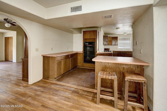 kitchen featuring light hardwood / wood-style flooring, a breakfast bar, ceiling fan, black double oven, and kitchen peninsula