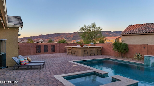 pool at dusk with an in ground hot tub, a mountain view, an outdoor bar, and a patio