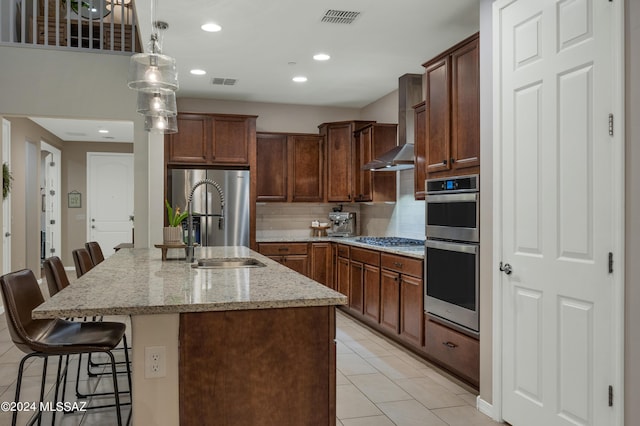 kitchen with appliances with stainless steel finishes, light stone counters, an island with sink, a kitchen bar, and wall chimney exhaust hood