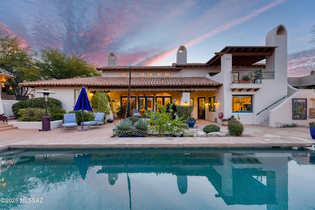 back of property at dusk featuring a balcony, a patio area, an outdoor pool, and a tiled roof