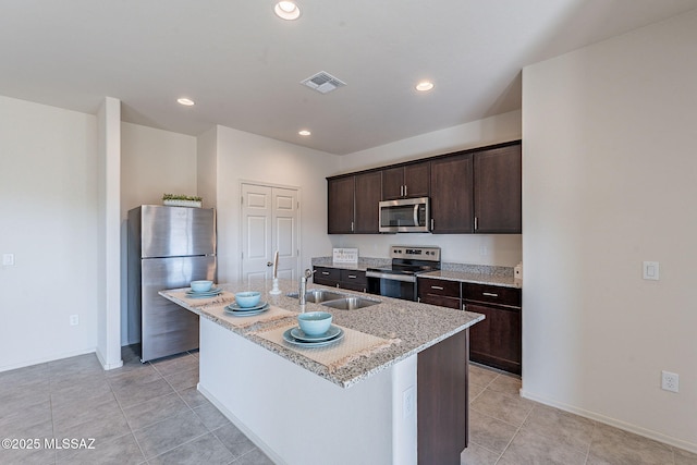 kitchen with an island with sink, sink, stainless steel appliances, light stone countertops, and dark brown cabinets