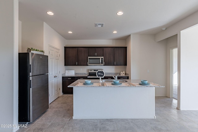 kitchen featuring light tile patterned floors, appliances with stainless steel finishes, a kitchen island with sink, dark brown cabinets, and light stone countertops