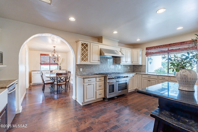 kitchen with arched walkways, dark wood-style flooring, wall chimney exhaust hood, and appliances with stainless steel finishes