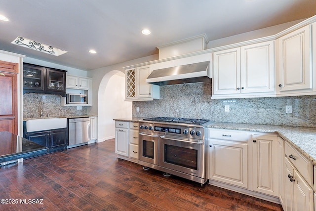 kitchen featuring wall chimney exhaust hood, appliances with stainless steel finishes, arched walkways, a sink, and dark wood-style floors