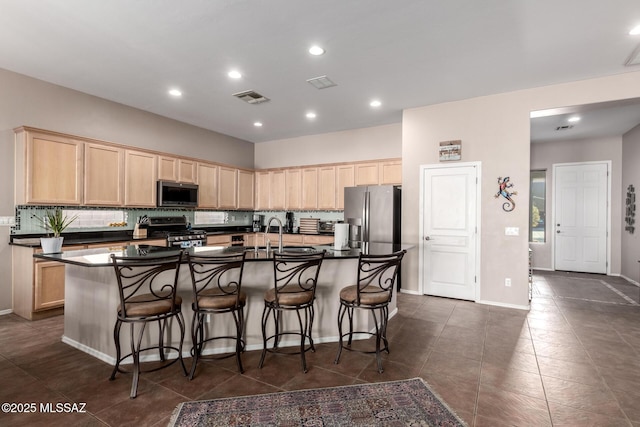 kitchen featuring dark tile patterned floors, stainless steel appliances, tasteful backsplash, an island with sink, and light brown cabinetry