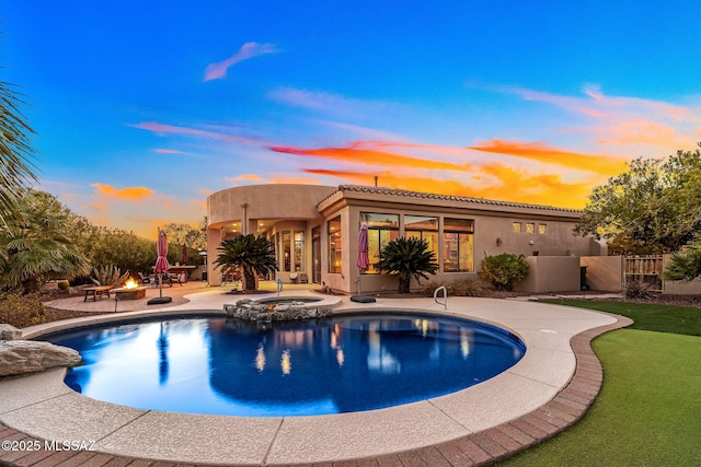 pool at dusk with a fire pit, a patio area, fence, and a pool with connected hot tub