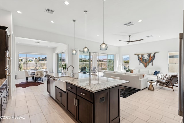 kitchen featuring dark brown cabinetry, sink, hanging light fixtures, a center island with sink, and light stone countertops