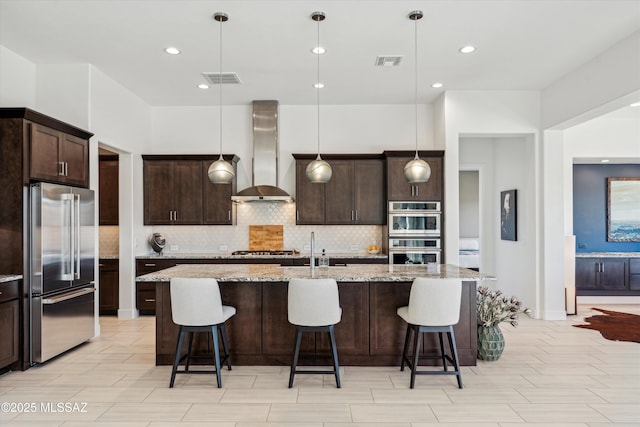 kitchen featuring light stone counters, decorative light fixtures, a center island with sink, stainless steel appliances, and wall chimney range hood