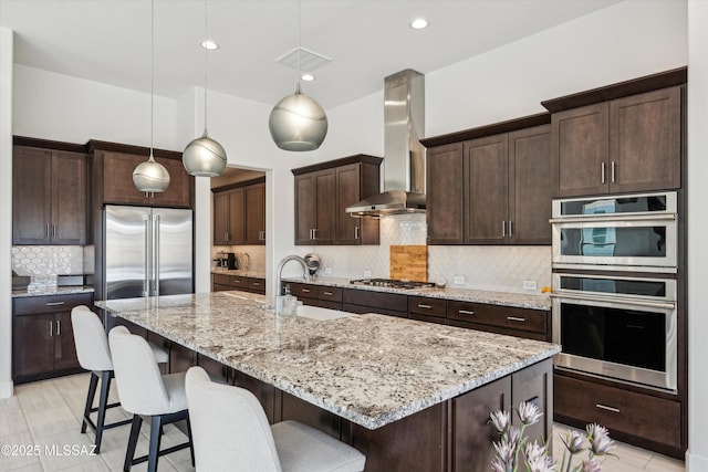 kitchen with wall chimney exhaust hood, decorative light fixtures, dark brown cabinets, a center island with sink, and appliances with stainless steel finishes