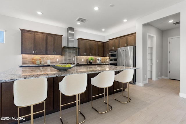 kitchen with dark brown cabinetry, wall chimney exhaust hood, high end refrigerator, a large island with sink, and stone counters