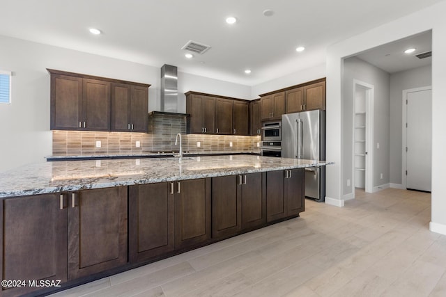 kitchen with decorative backsplash, dark brown cabinetry, light stone countertops, high end fridge, and wall chimney range hood