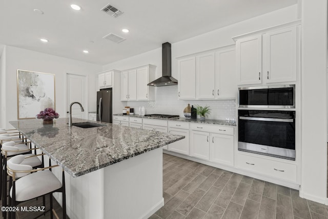 kitchen featuring wall chimney range hood, sink, a kitchen island with sink, stainless steel appliances, and white cabinets