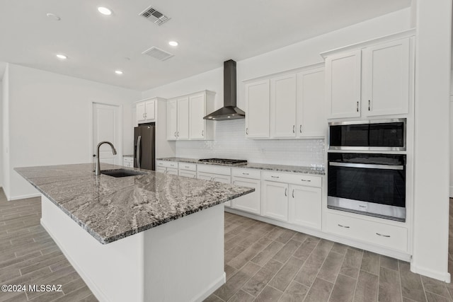 kitchen featuring wall chimney exhaust hood, sink, appliances with stainless steel finishes, an island with sink, and white cabinets