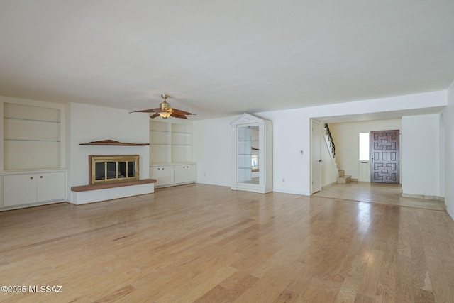 unfurnished living room with built in features, light wood finished floors, a ceiling fan, a glass covered fireplace, and stairs