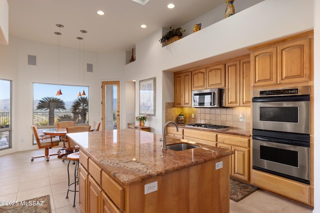 kitchen with an island with sink, light stone counters, appliances with stainless steel finishes, pendant lighting, and a sink