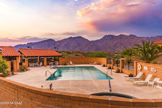 pool at dusk featuring a jacuzzi, fence, a mountain view, a community pool, and a patio area