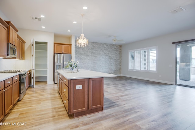 kitchen featuring appliances with stainless steel finishes, decorative light fixtures, an island with sink, sink, and light hardwood / wood-style flooring