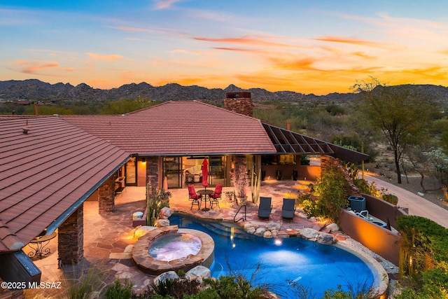 pool at dusk with an in ground hot tub, a mountain view, and a patio area