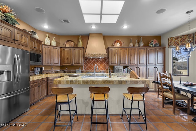 kitchen with stainless steel refrigerator with ice dispenser, built in microwave, sink, a center island with sink, and custom range hood