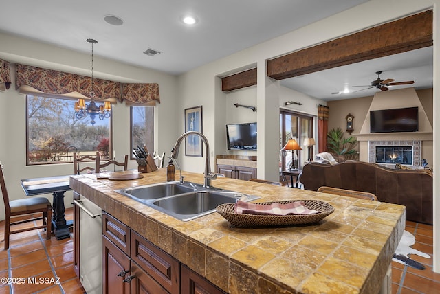 kitchen featuring sink, hanging light fixtures, a center island with sink, dishwasher, and a tile fireplace