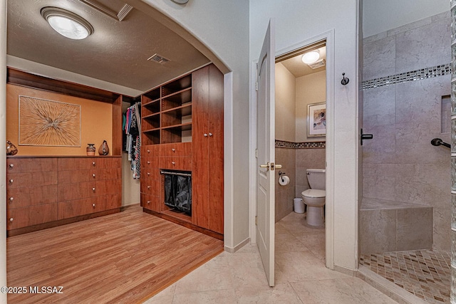 bathroom featuring toilet, wood-type flooring, tile walls, a tile shower, and a textured ceiling