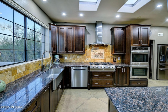 kitchen with backsplash, appliances with stainless steel finishes, dark brown cabinetry, a sink, and wall chimney exhaust hood