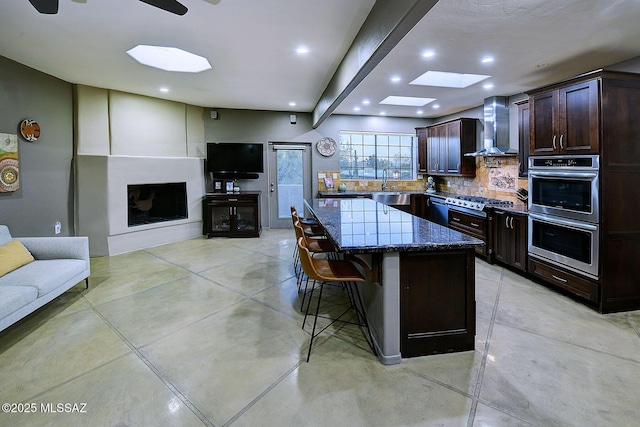 kitchen featuring dark stone counters, a kitchen breakfast bar, dark brown cabinets, stainless steel double oven, and wall chimney range hood