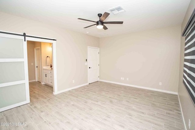 unfurnished bedroom with ensuite bathroom, a barn door, ceiling fan, and light wood-type flooring