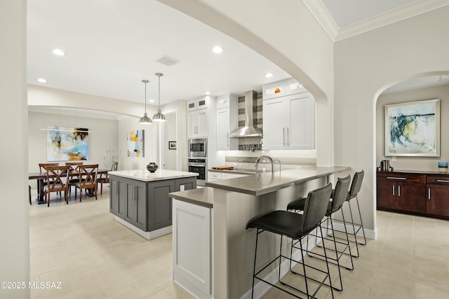 kitchen featuring appliances with stainless steel finishes, white cabinetry, hanging light fixtures, kitchen peninsula, and wall chimney exhaust hood