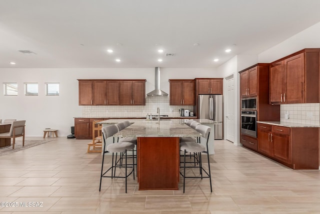 kitchen featuring appliances with stainless steel finishes, an island with sink, a breakfast bar area, light stone counters, and wall chimney range hood