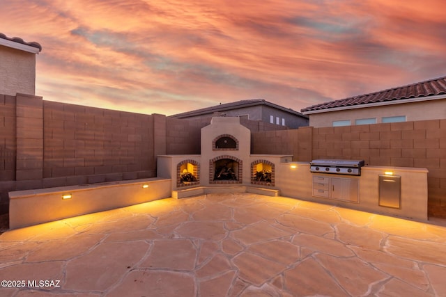 patio terrace at dusk featuring exterior kitchen, a grill, and exterior fireplace