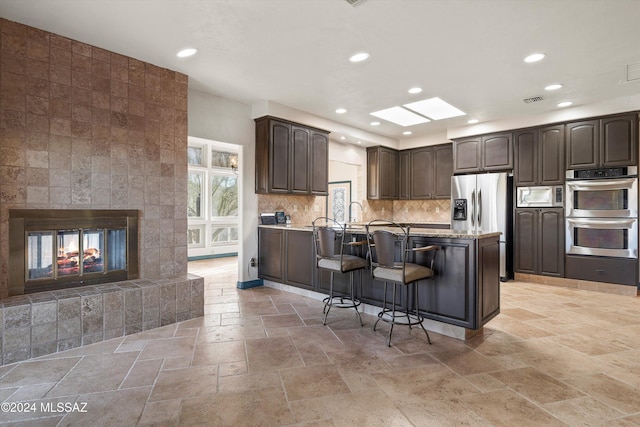 kitchen featuring a breakfast bar area, stone tile floors, stainless steel appliances, dark brown cabinetry, and a multi sided fireplace