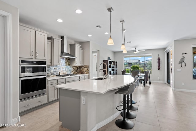 kitchen with a sink, gray cabinets, wall chimney exhaust hood, light countertops, and a center island with sink