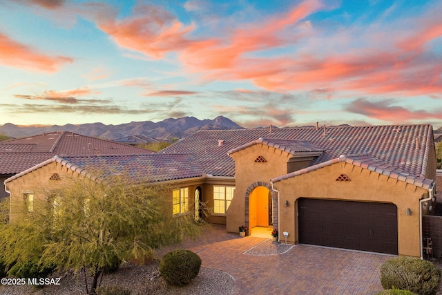 mediterranean / spanish-style house with an attached garage, a mountain view, a tiled roof, decorative driveway, and stucco siding