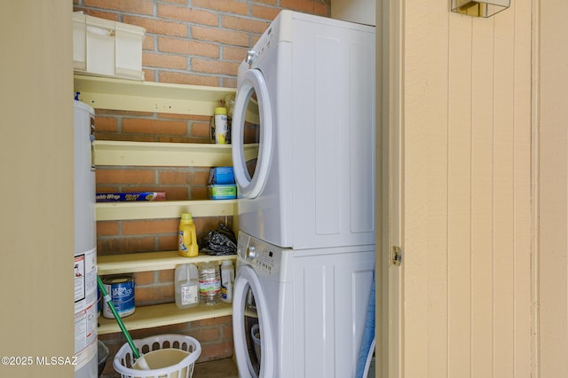 clothes washing area featuring water heater, brick wall, and stacked washing maching and dryer